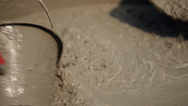 Wet cement pouring from scoop into container, close-up. Construction material mixing and pouring process for building, flooring, or foundation work.