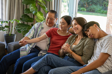 Diverse family sitting on gray upholstered sofa in living room holding hands near plant and door
