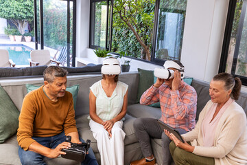 Four friends sitting on gray sectional sofa using white VR headsets, holding tablet plus controller