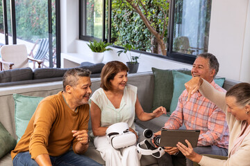 Friends including senior woman sitting on gray sofa at home sharing white VR headset and tablet