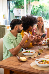 Diverse friends sitting at wooden dining table at home, sharing smartphone and bowls of pasta