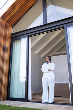 African American mid adult woman standing at glass door, holding phone in pale pajamas and slippers