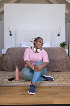 African American female sitting on woven rug by bed, wearing pink top, teal leggings, holding phone