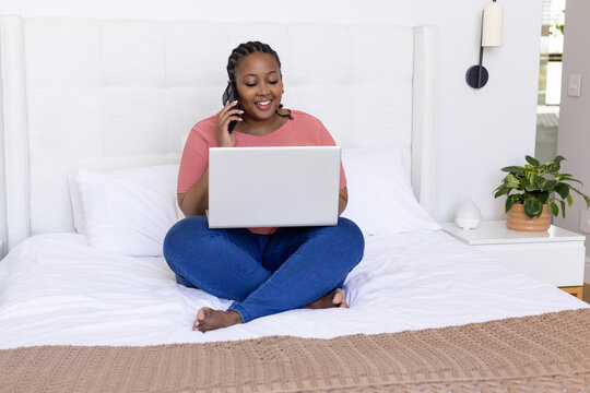 Mid-adult African American woman sitting cross-legged on bed, using laptop and talking on phone