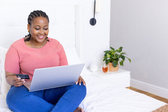African American woman wearing coral-pink top and jeans on white bed using laptop with credit card