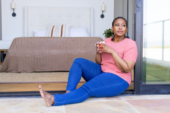 Mid adult African American woman sitting on threshold between bedroom and patio holding glass mug