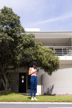 African American woman standing beneath tree on lawn checking smartphone, wearing sunglasses