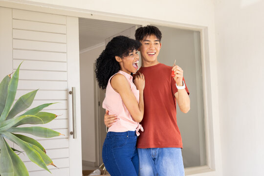 Diverse couple in their twenties in jeans standing at front doorway celebrating, man holding key