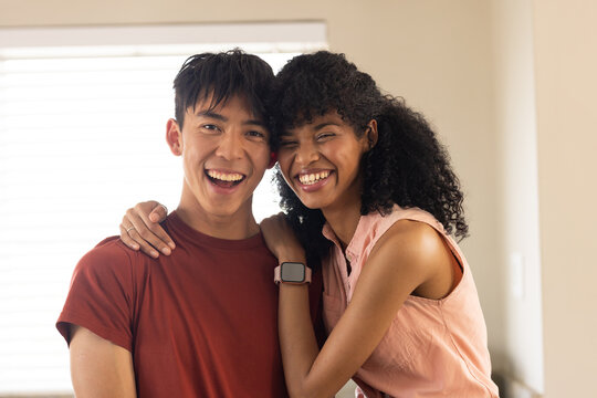 Diverse couple smiling and leaning near horizontal blinds, wearing casual clothes with smartwatch