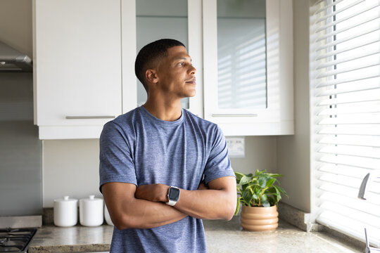 African American man standing arms crossed, wearing blue tee gazing through blinds at counter plant