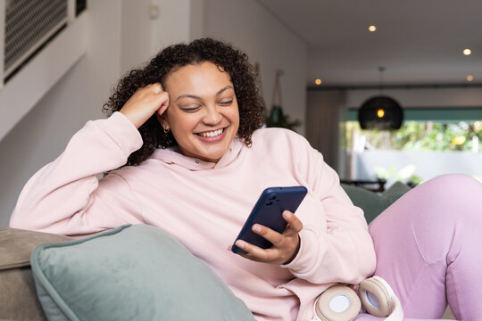 African-American woman reclining on sofa at home, wearing pink hoodie, holding blue phone