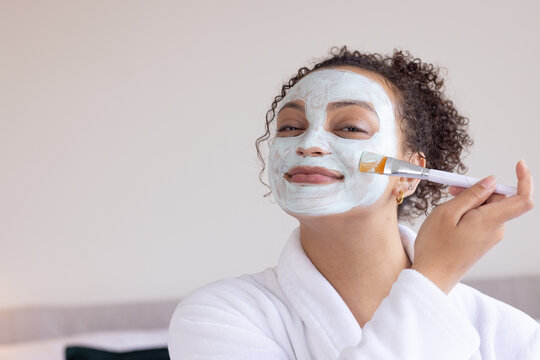 African woman sitting in home spa, wearing white bathrobe, applying green face mask with flat brush