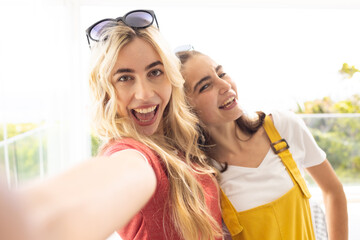 Female friends taking selfie on balcony, holding phone, in red top and yellow overalls