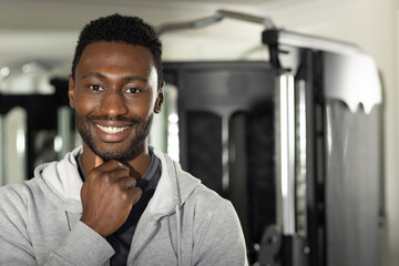 Mid adult African American man smiling, resting chin in gym wearing gray hoodie with cable machine