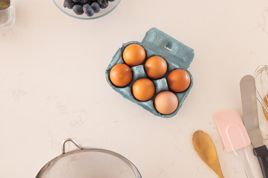 Open teal carton of six brown eggs is sitting on beige countertop, near spoon and whisk