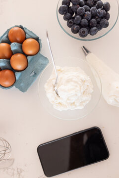 Glass bowl of whipped cream is sitting on light countertop, with egg carton, piping bag, smartphone