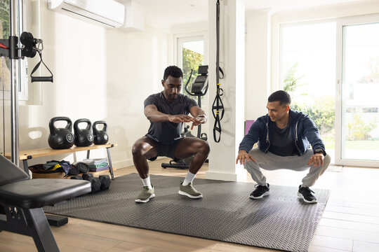 Diverse male workout partners performing squats cueing form on mat at home gym with kettlebells