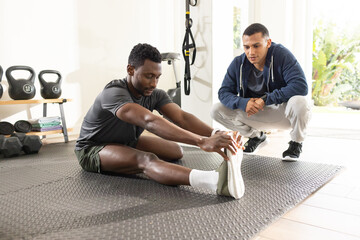 Male client stretching in home gym on rubber mat, African-American trainer coaching with kettlebell