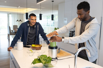 Male friends preparing green smoothie at modern kitchen island using blender and leafy greens