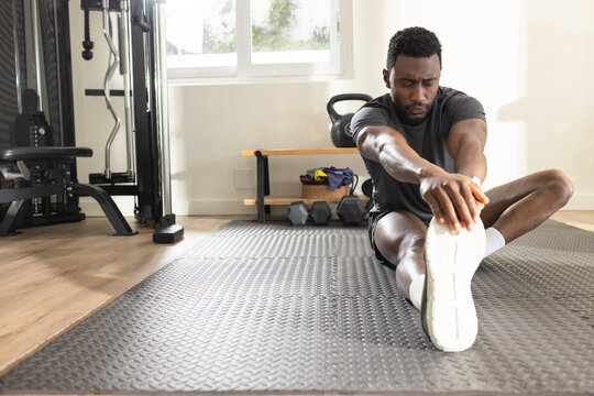 African American man stretching on mat in home gym in sportswear, kettlebell nearby, copy space