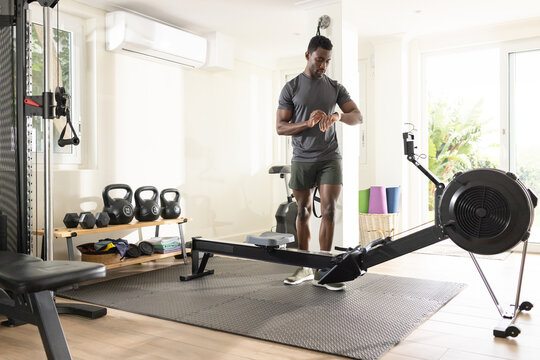 African American man wearing gray shirt, olive shorts checking watch in home gym by rower