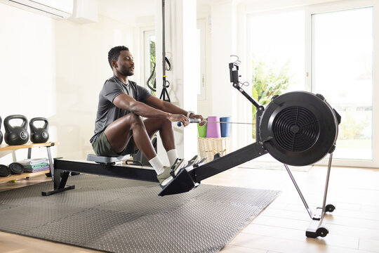 African American man in mid thirties rowing on rowing machine in home gym, kettlebells visible