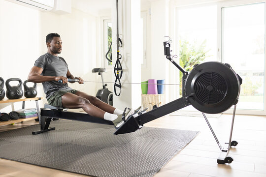 African American man rowing on rowing machine in home gym wearing grey tee, green shorts