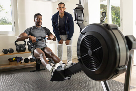 Diverse client in grey tee pulling on rowing machine flywheel while trainer coaching near bench