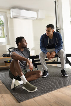 Diverse male friends resting and talking in small home gym with bench, TRX strap, dumbbells
