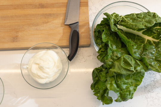Swiss chard is spilling from glass bowl onto kitchen countertop, knife on board, yogurt bowl nearby