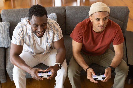 Diverse male friends playing console games on gray couch in living room holding white controllers