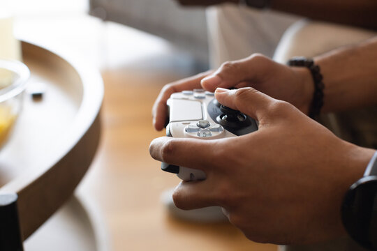Hands are holding white wireless game controller in living room, wooden side table with glass bowl