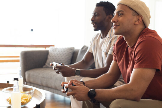 Male friends gaming on home couch, holding controllers by chips bowl, wearing beanie, striped shirt