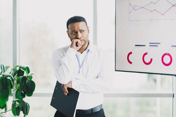 Thoughtful businessman stands in a bright office with charts planning strategy during a busy meeting