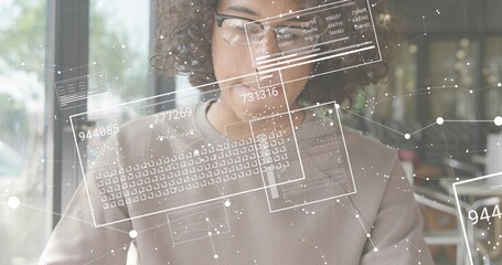 Leaning man wearing knit sweater, glasses scanning laptop with data overlay by cafe window plants