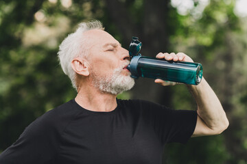 Elderly bearded man in athletic wear drinks from a bottle outdoors in a park enjoying a refreshing moment during a sunny day