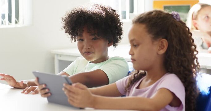 African American children at desk right child holding tablet tapping left child speaking for class
