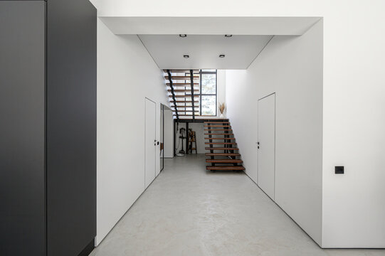 Minimalist hallway with white walls, light grey floor, dark grey cabinet, wooden staircase with open risers, recessed ceiling lights, large window, clean lines, modern aesthetic.