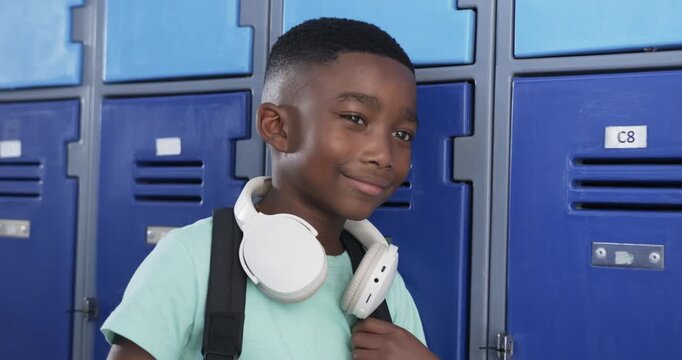 Teenage boy noticing camera, adjusting backpack strap, smiling with headphones at school lockers C8