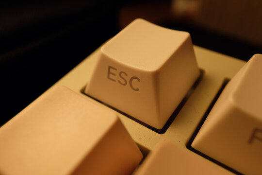 Macro view of a vintage beige mechanical keyboard focusing on the Esc key. Retro computing technology with warm lighting emphasizing the textured plastic surface and classic font.
