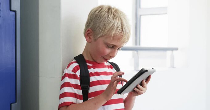 School-age boy leaning in hall, tapping tablet while viewing content, smiling, striped tee backpack