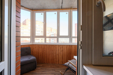 Fototapeta premium Balcony with wooden paneling, large panoramic windows, dark upholstered sofa, folding chair, brick wall section, wooden flooring, cozy and bright space.