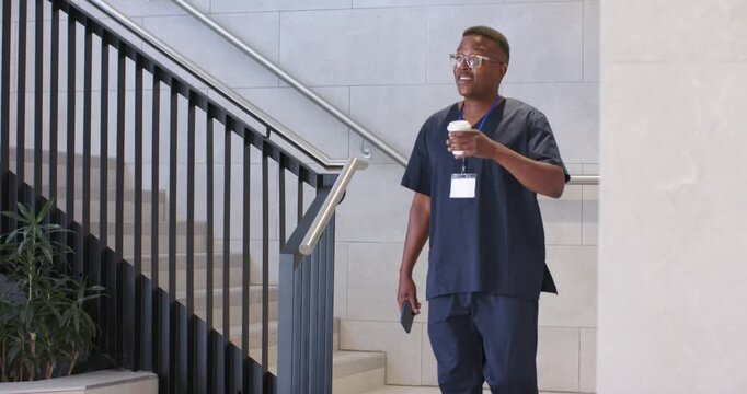 Male coworkers in navy scrubs descending stairs in lobby with coffee cups, smartphone taking break