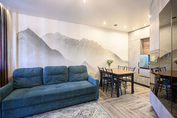 Cozy living-dining area with teal sofa, mountain mural wall, wooden dining set, light wood flooring, modern kitchen cabinetry, patterned rug, sheer curtains, ambient lighting. © evgeniykleymenov