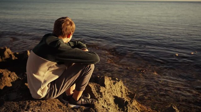 Thoughtful preteen boy sitting by the sea during sunset. Child enjoying quiet moment near ocean. Concept of childhood, emotions, mental health, calm lifestyle, freedom, nature and peaceful solitude.