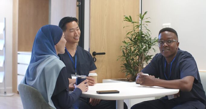 Diverse coworkers taking break in breakroom in navy scrubs chatting, holding coffee, IDs on table