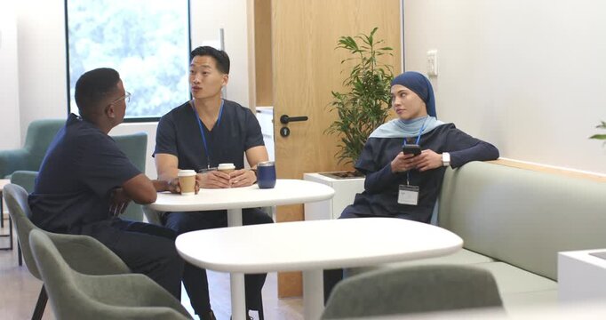 Diverse staff in scrubs on break at lounge, center with coffee starting talk, woman checking phone