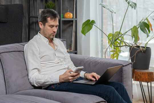 Man checking smartphone and using laptop while sitting on sofa in living room. Freelancer multitasking with mobile phone and computer at home office. Remote worker using digital devices online.