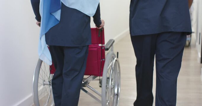 Diverse staff in scrubs escorting red wheelchair, walking hospital hall, tablet, stethoscope, hijab