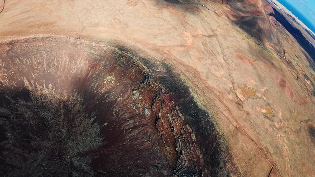 Aerial drone sweeps over volcanic craters and cinder cones in Fuerteventura, showing black interiors, sandy plains, lava rims, vents, tracks, and distant coastline.